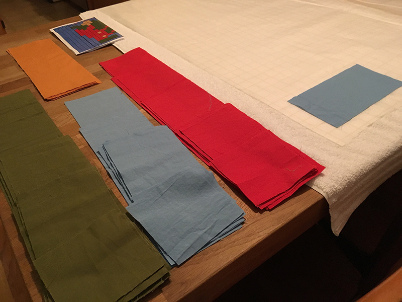Pre-cut cloth squares in red, blue, green, and yellow-brown lie waiting for placement on the grid interfacing.